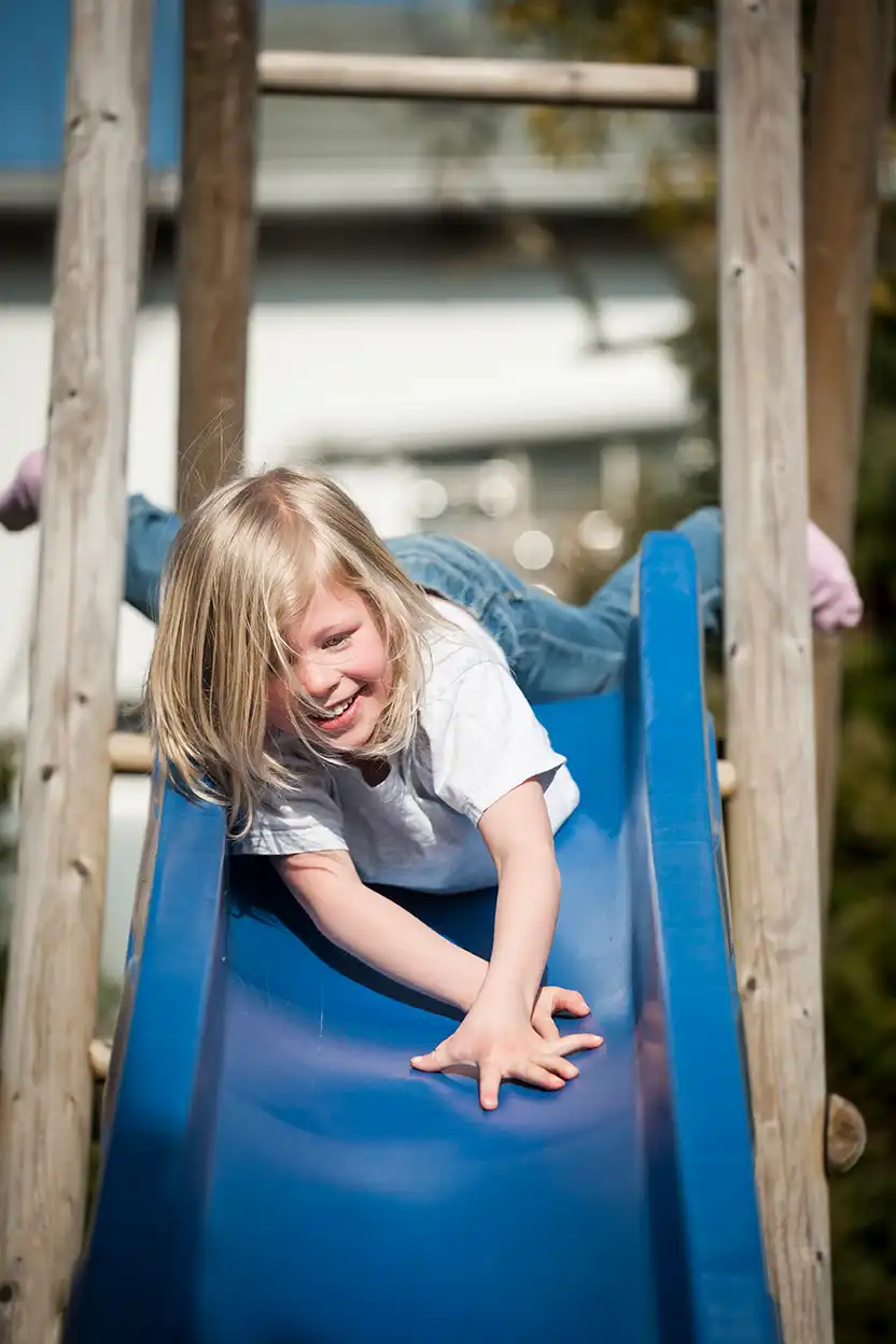 Ein blondhaariges Mädchen rutscht lachend auf einer blauen Rutsche eines Spielturms
